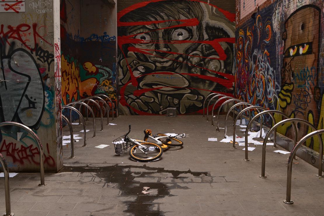 An oBike lies buckled on the ground amongst ample docking bays, while graffitied characters look on.